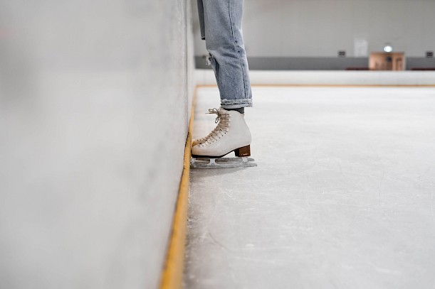 Rand einer Eisbahn, an der eine Person steht, von der nur die Beine und die weißen Schnittschuhe zu sehen sind.